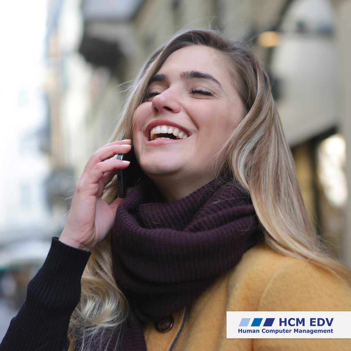 Blonde Frau mit gelber Jacke und lila Schal telefoniert lachend mit dem Smartphone.