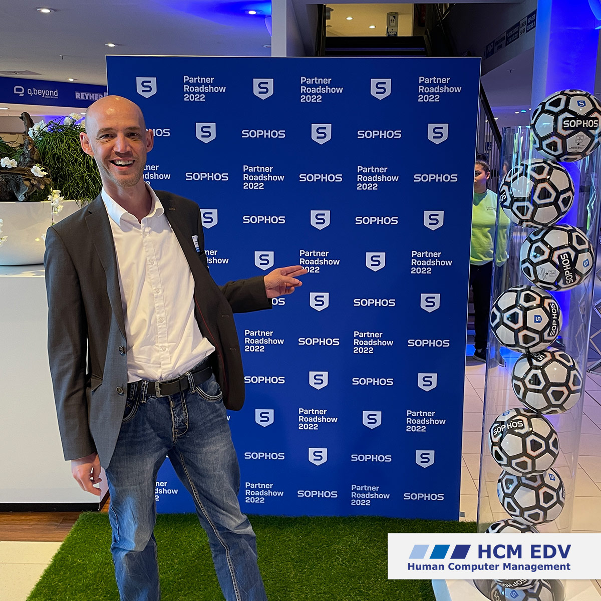 Thomas Buss von der HCM EDV in grauem Jacket und Jeans vor einer blau-weißen Werbewand der Sophos Road Show 2022.